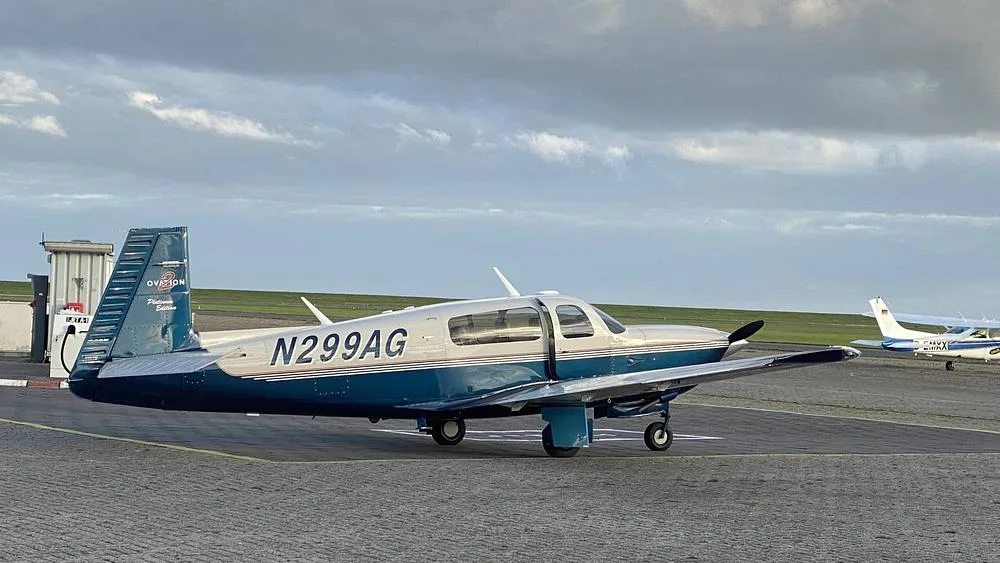 A Mooney Ovation 2 Platinum Edition aircraft, N299AG, with a blue and silver paint scheme, parked on a tarmac under a cloudy sky.