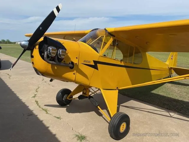 A side-front view of a yellow Piper Cub-style aircraft with a black propeller, parked on a concrete pad.