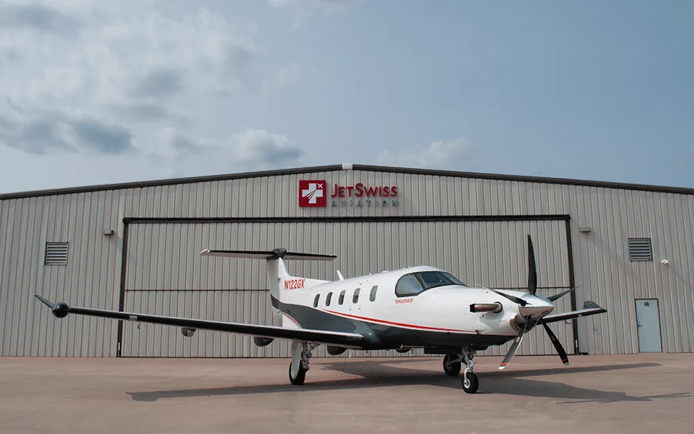 A white and grey Pilatus PC-12 turboprop aircraft with tail number N122GX parked on the tarmac in front of a hangar under a cloudy sky.