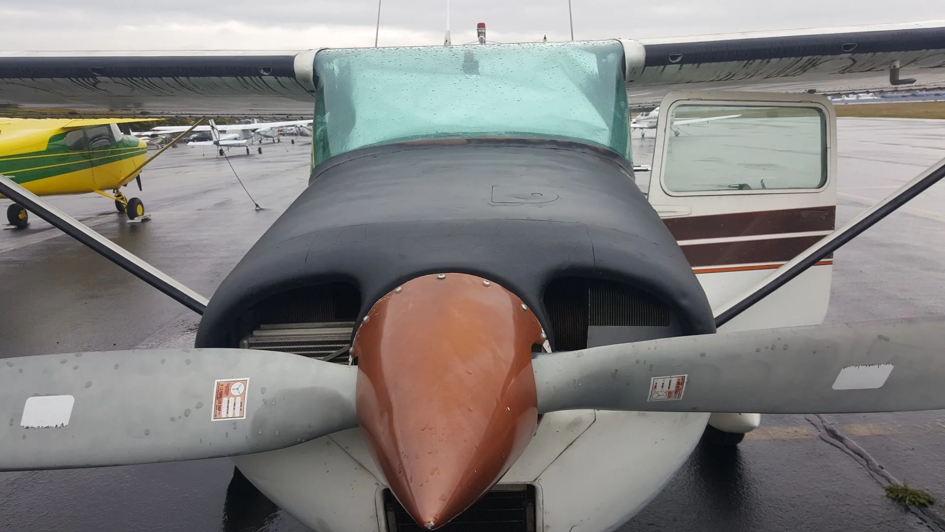 A close-up front view of a white and brown Cessna 182 aircraft on a wet tarmac, with its propeller and engine cowling visible.