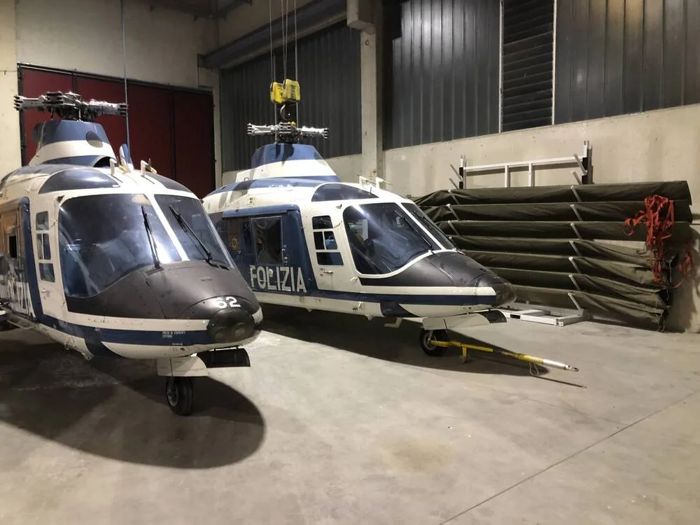 Two blue and white police helicopters, marked "POLIZIA", are parked in a hangar with equipment in the background.