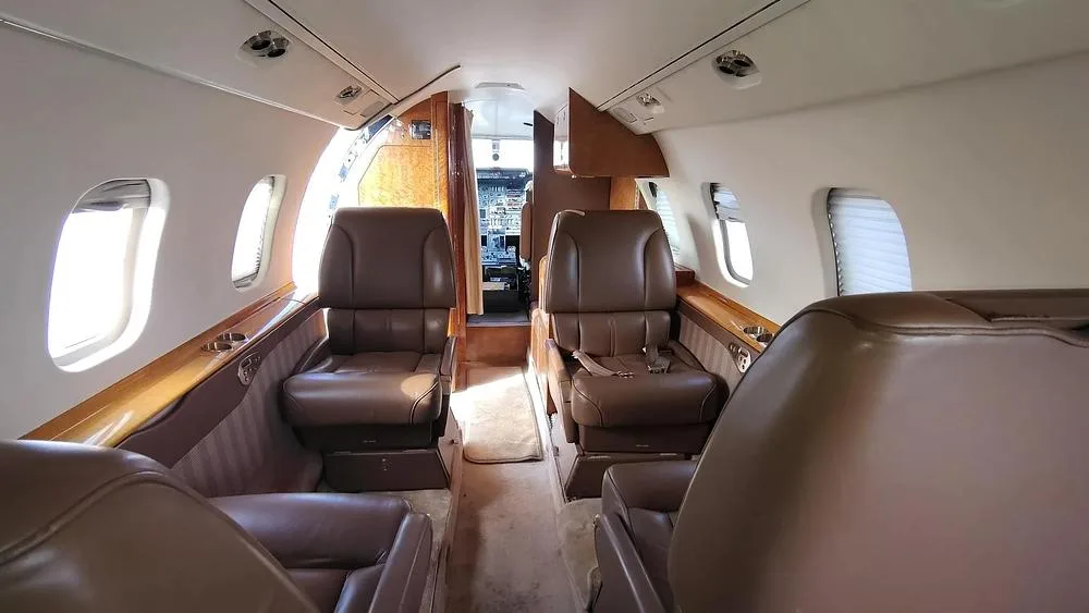 The interior cabin of a private jet with brown leather seats and wood trim, looking forward towards the cockpit.