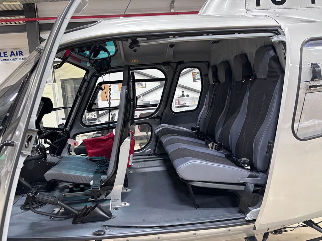 The interior cabin of a silver Airbus H125 helicopter, showing the pilot's seat and passenger bench seats.