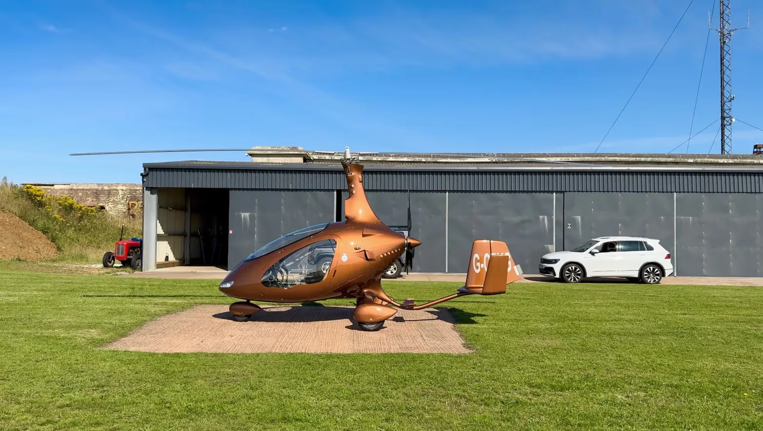 A brown gyrocopter, G-ORYG, parked on a concrete pad in front of a hangar with a white SUV.