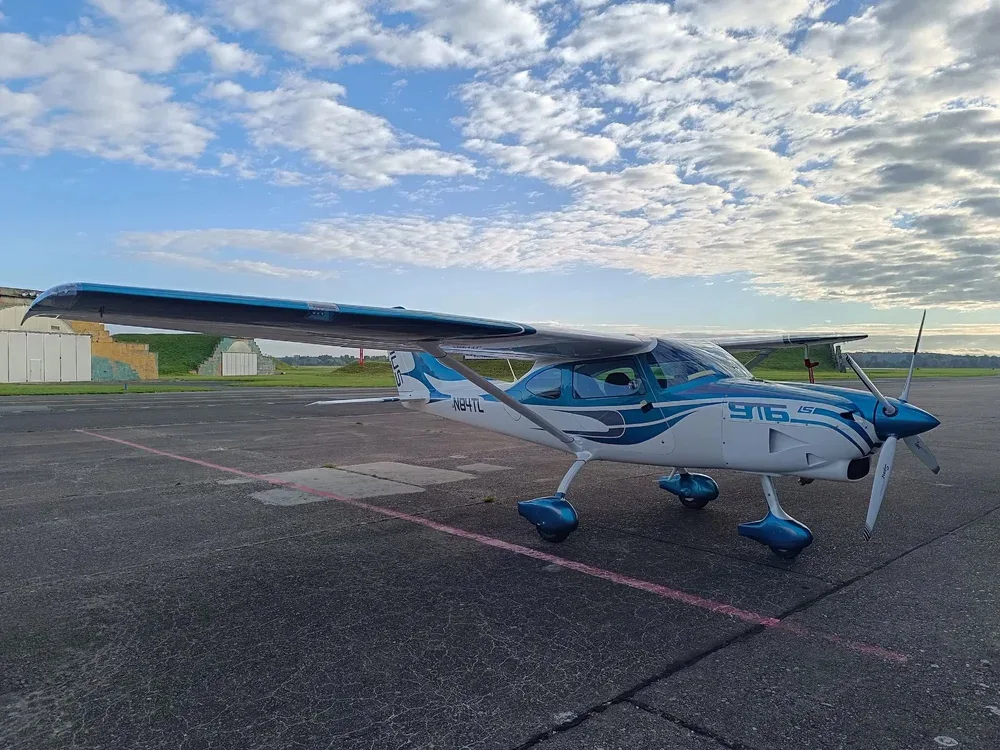 A white and teal Sirius 916 LS aircraft, registration N84TL, parked on an asphalt tarmac with a clear horizon under a partly cloudy sky.
