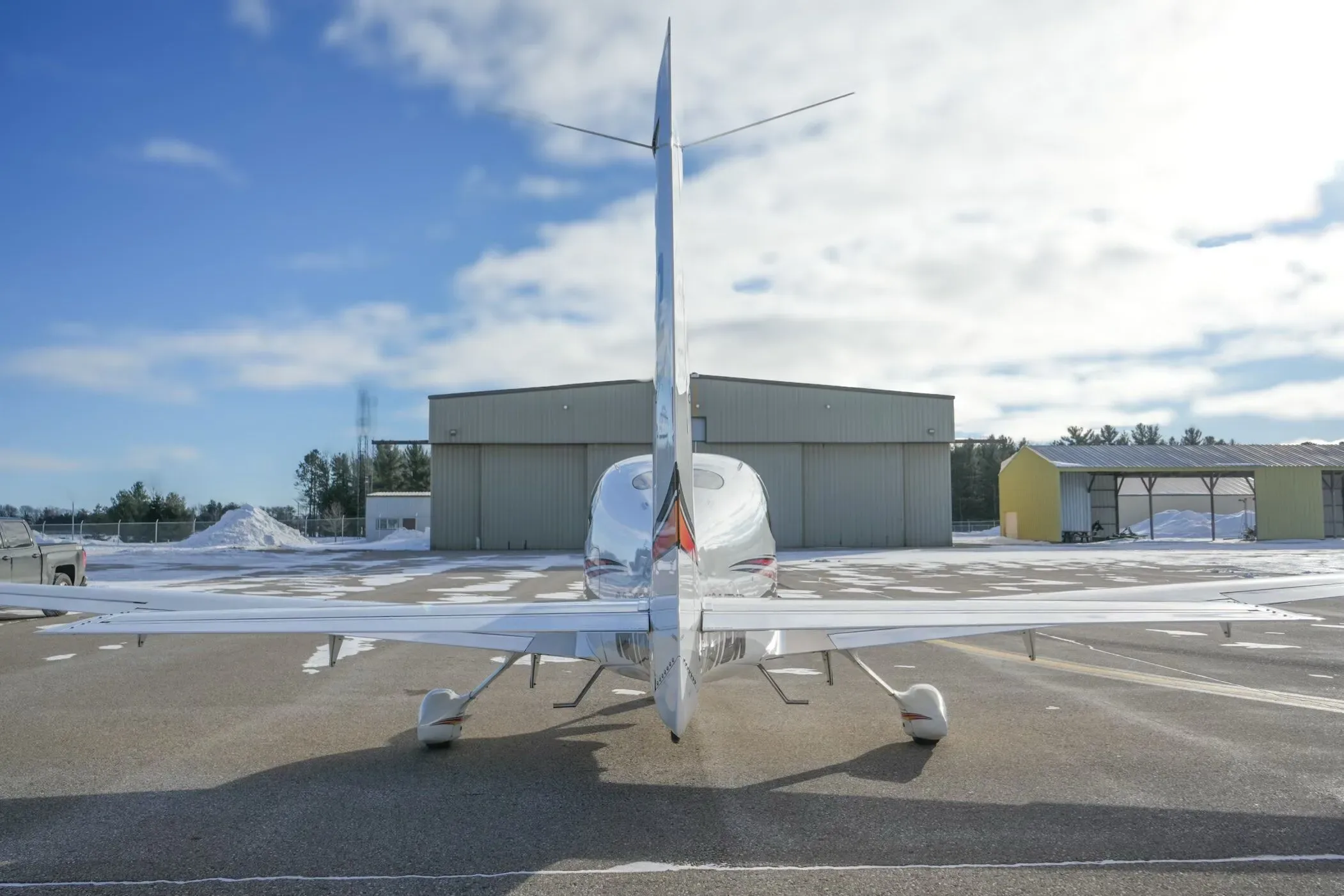 A rear view of a white Cirrus SR22 aircraft parked on a snowy airfield with hangars under a blue sky.