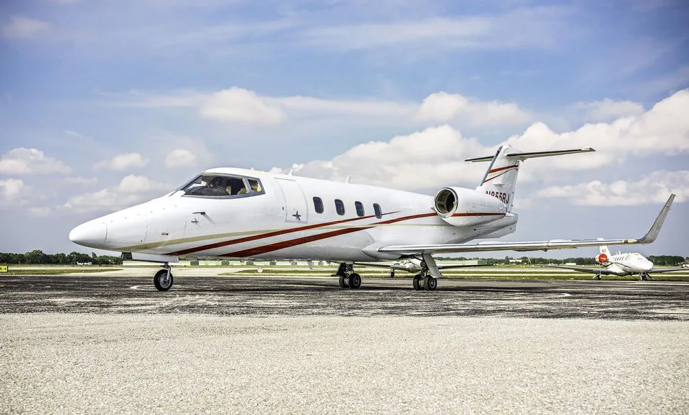 A white Learjet 45 with red and gold stripes, registration N855RJ, parked on an airport tarmac under a cloudy sky.