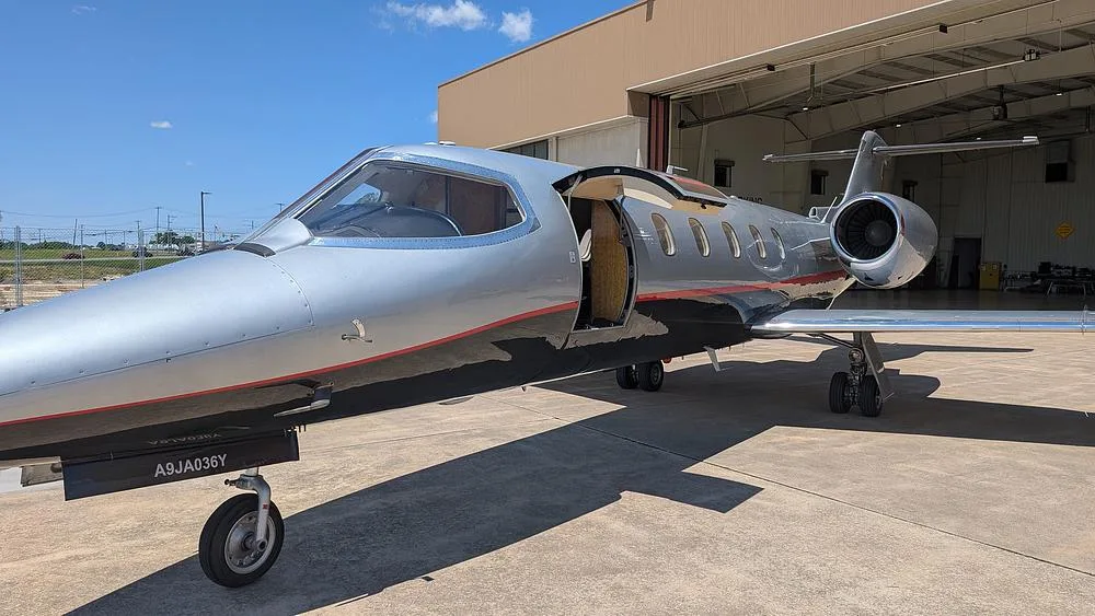 A silver and black Learjet with a red stripe, parked on a tarmac with its main cabin door open, in front of a hangar.