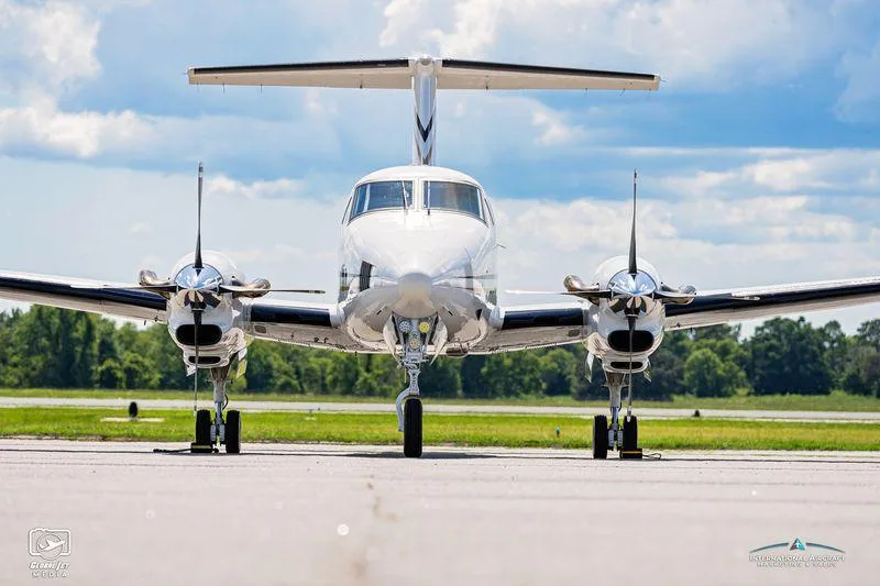 Front view of a white twin-engine turboprop aircraft with black and gold accents on a tarmac under a blue sky.
