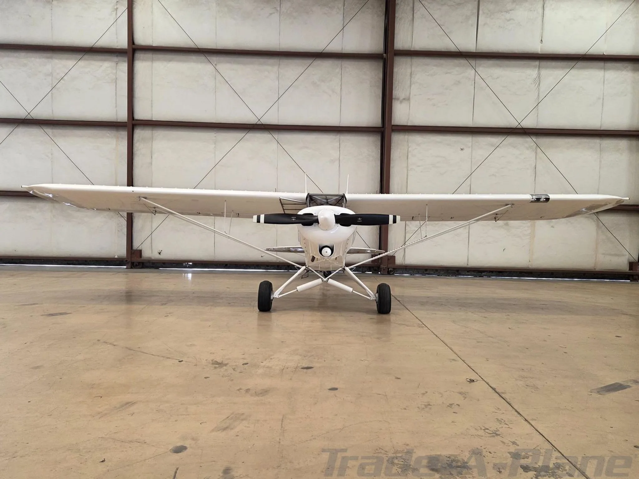 Front view of a white Piper Super Cub aircraft with a black propeller, parked in a hangar.