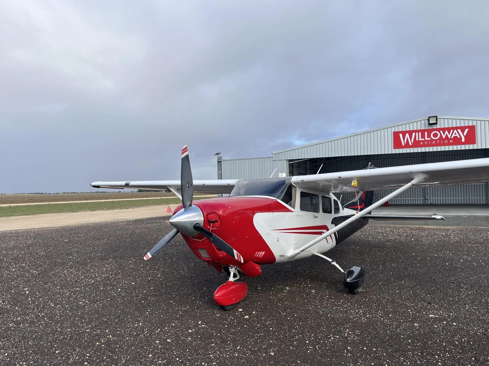 A red and white single-engine propeller aircraft is parked on an airfield in front of a WilloWay Aviation hangar under an overcast sky.