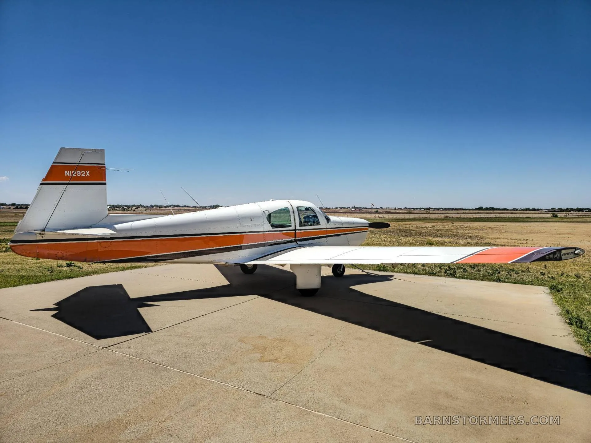 A white and orange single-engine aircraft (N282X) parked on a concrete pad, viewed from the side under a clear blue sky.