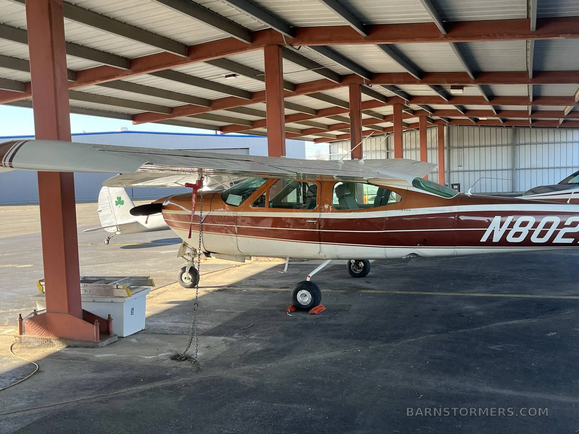 A brown and white single-engine propeller aircraft with tail number N8022G is parked under a metal hangar structure.