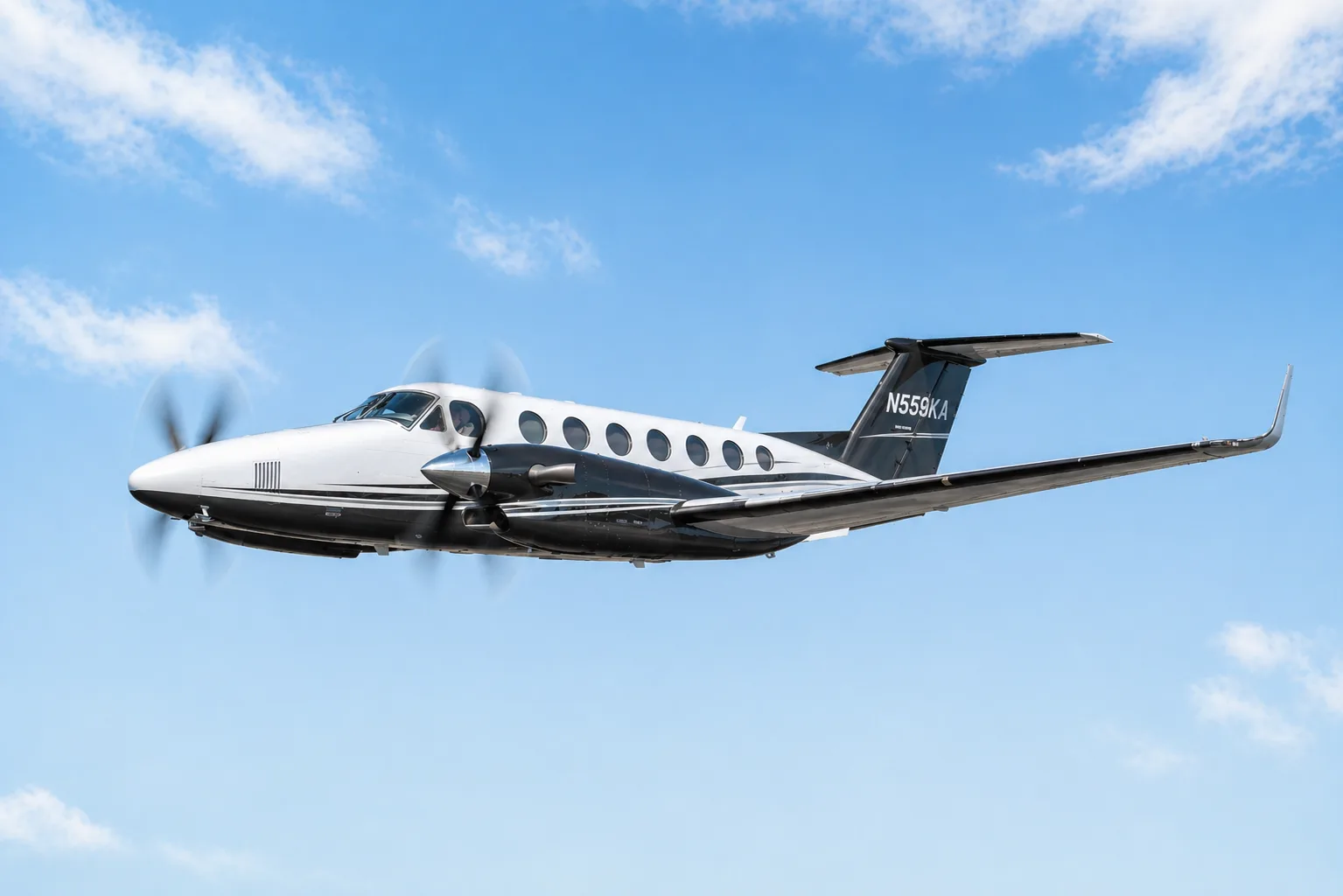 A white and black King Air 350i aircraft with registration N559KA flying against a blue sky with clouds.