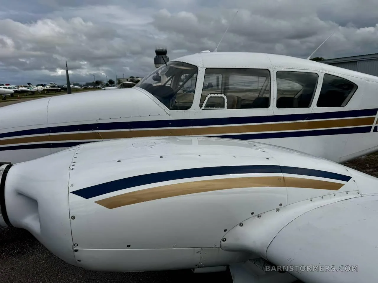 A close-up side view of a white twin-engine aircraft with blue and gold stripes, showing the cockpit and engine cowling, with an airport tower in the background.