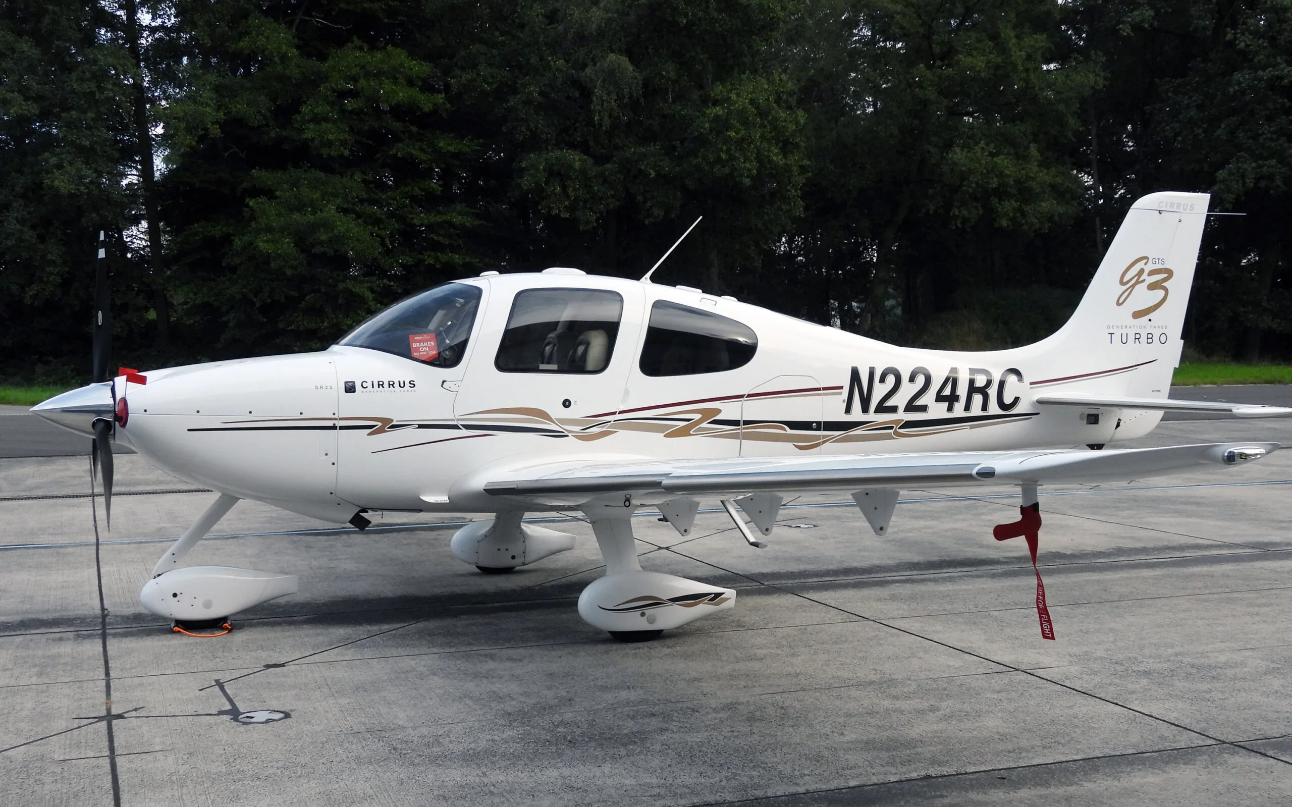 A white Cirrus SR22T (N224RC) with brown and black stripes parked on an asphalt apron with trees in the background.
