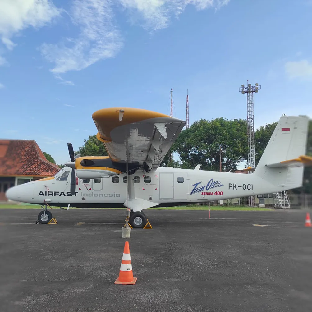 A side view of a white and yellow Airfast Indonesia Twin Otter Series 400 aircraft (PK-OCI) with its wing flaps extended, parked on an airfield with buildings and towers in the background.