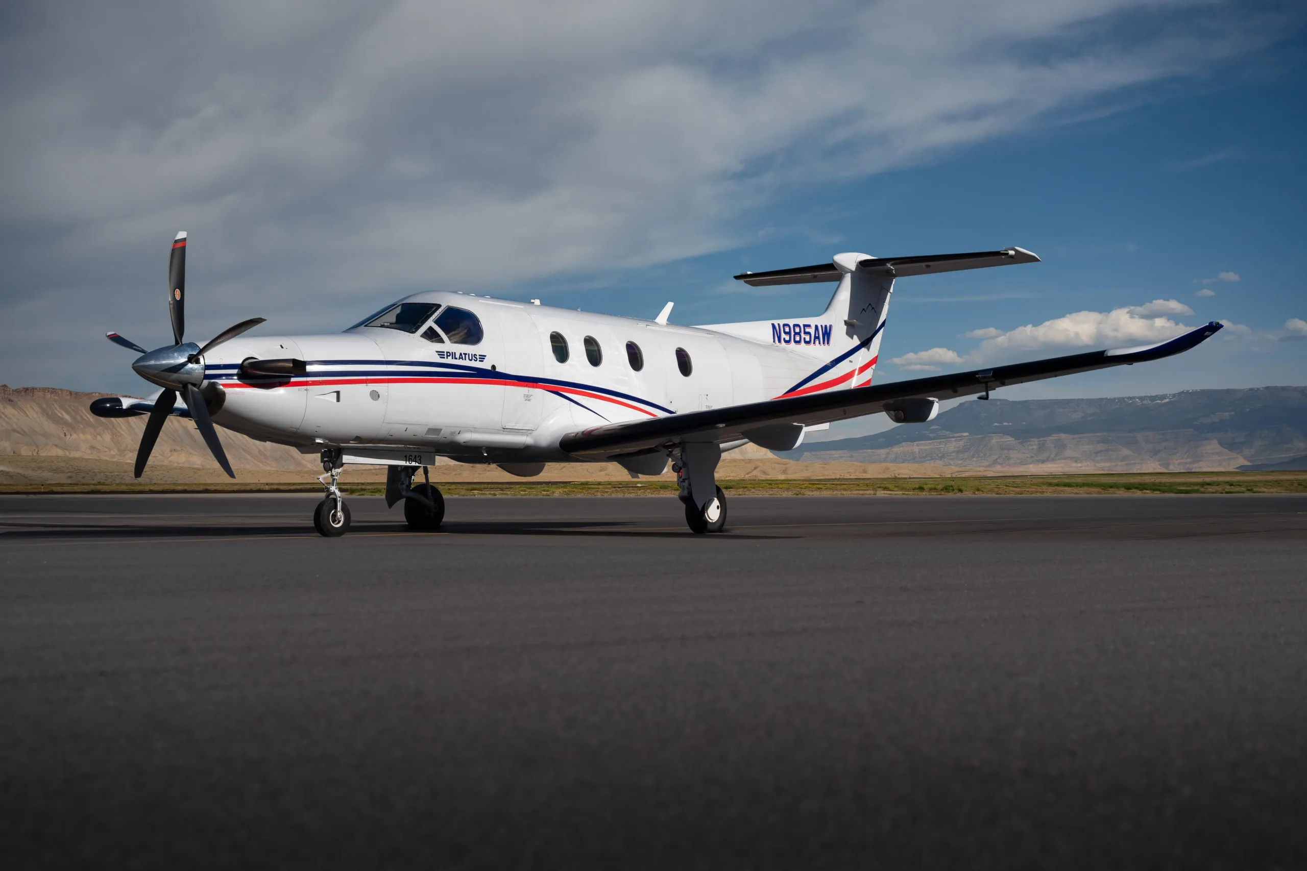 A Pilatus PC-12 aircraft, N985AW, with a white, blue, and red paint scheme, sits on a tarmac with mountains in the background.