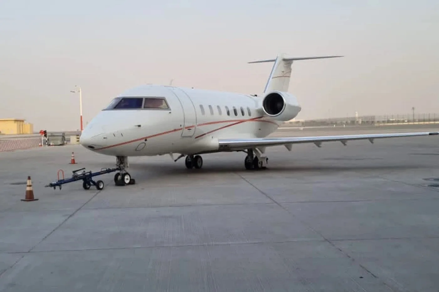 A white business jet with red pinstripes parked on an airport tarmac with ground equipment nearby.