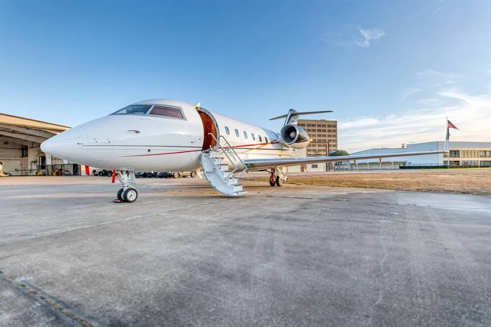 A white business jet with its main door open and stairs extended, parked on an airfield with hangars and a building in the background.