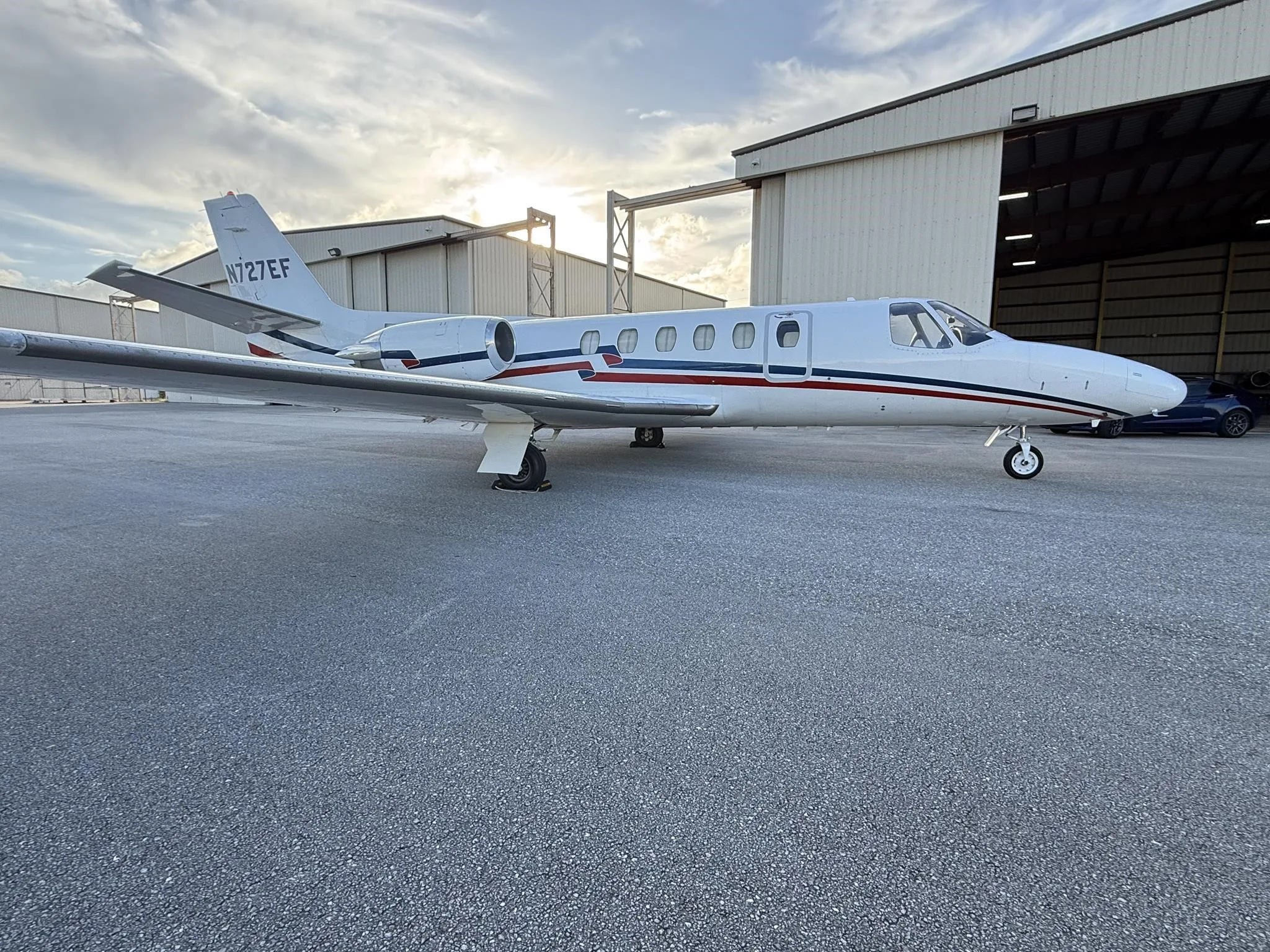 A white Cessna Citation business jet (N727EF) with red and blue stripes parked on an airfield at sunset.