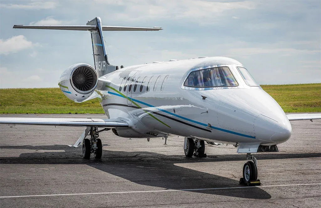 A white business jet with blue and green stripes and registration N545XR on its tail, parked on a tarmac with a grassy field in the background.