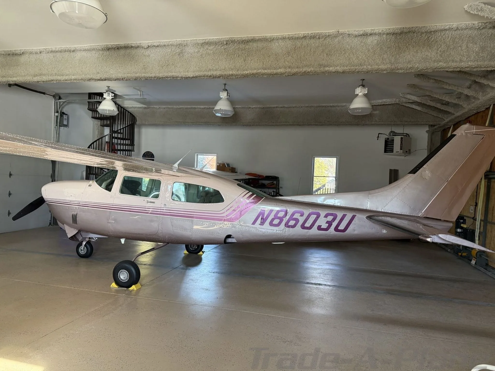 A side view of a light pink and purple Cessna 210 Centurion aircraft, registration N8603U, parked in a hangar.