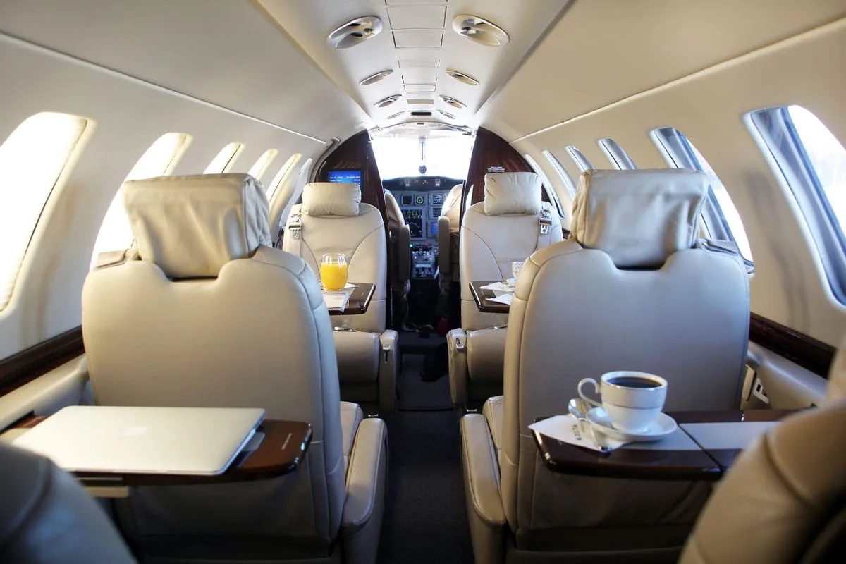 The interior cabin of a private jet with beige leather seats, fold-out tables, a laptop, and a cup of coffee.