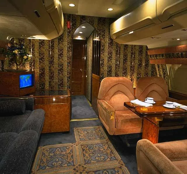 Another view of the luxurious aircraft interior with brown velvet seats, a small table, a television, and patterned wallpaper.