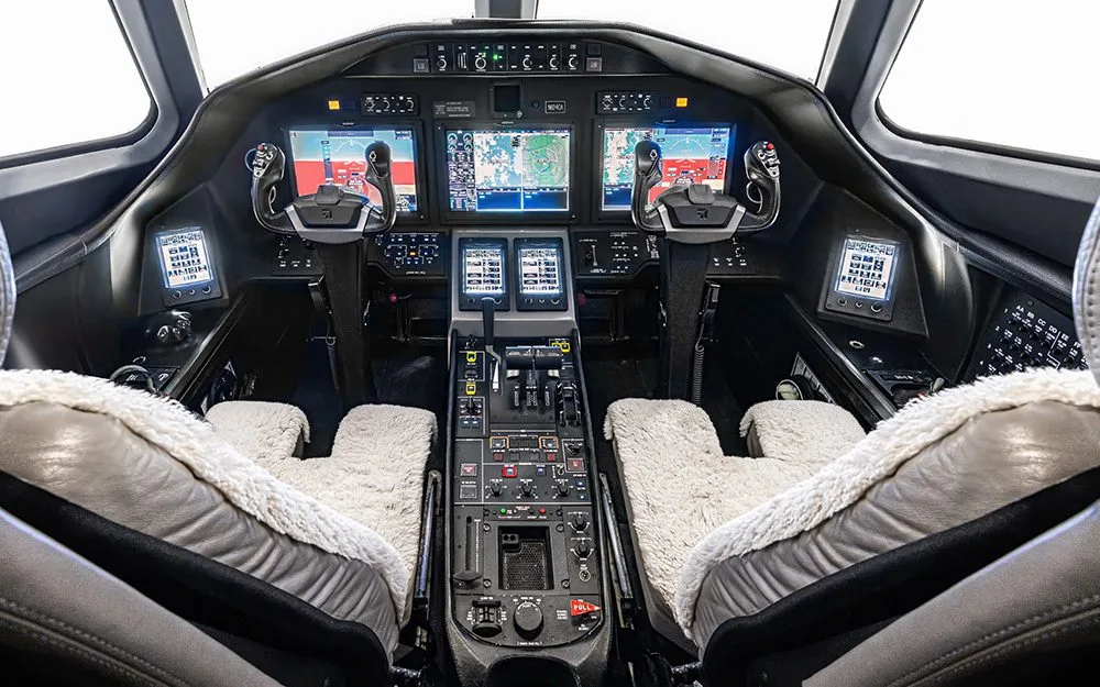 The modern glass cockpit of a business jet, featuring multiple digital displays and two pilot seats with sheepskin covers.