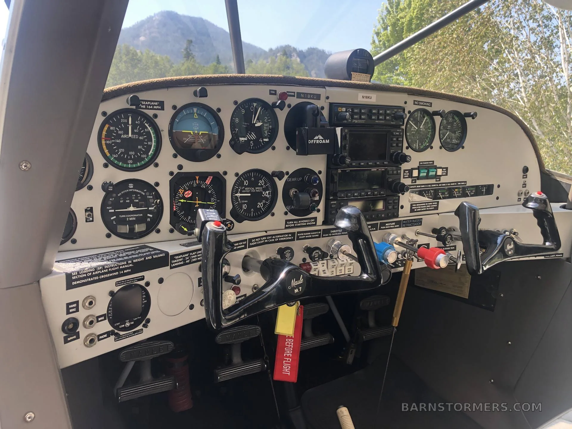 The cockpit of a floatplane, showing the instrument panel, yoke, and controls with mountains visible through the windshield.
