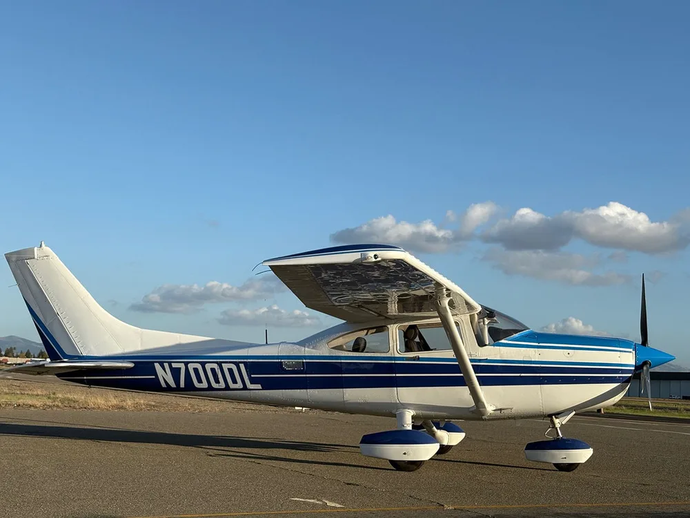 A white and blue Cessna 182 Skylane (N700DL) parked on an airfield, viewed from the front-right.