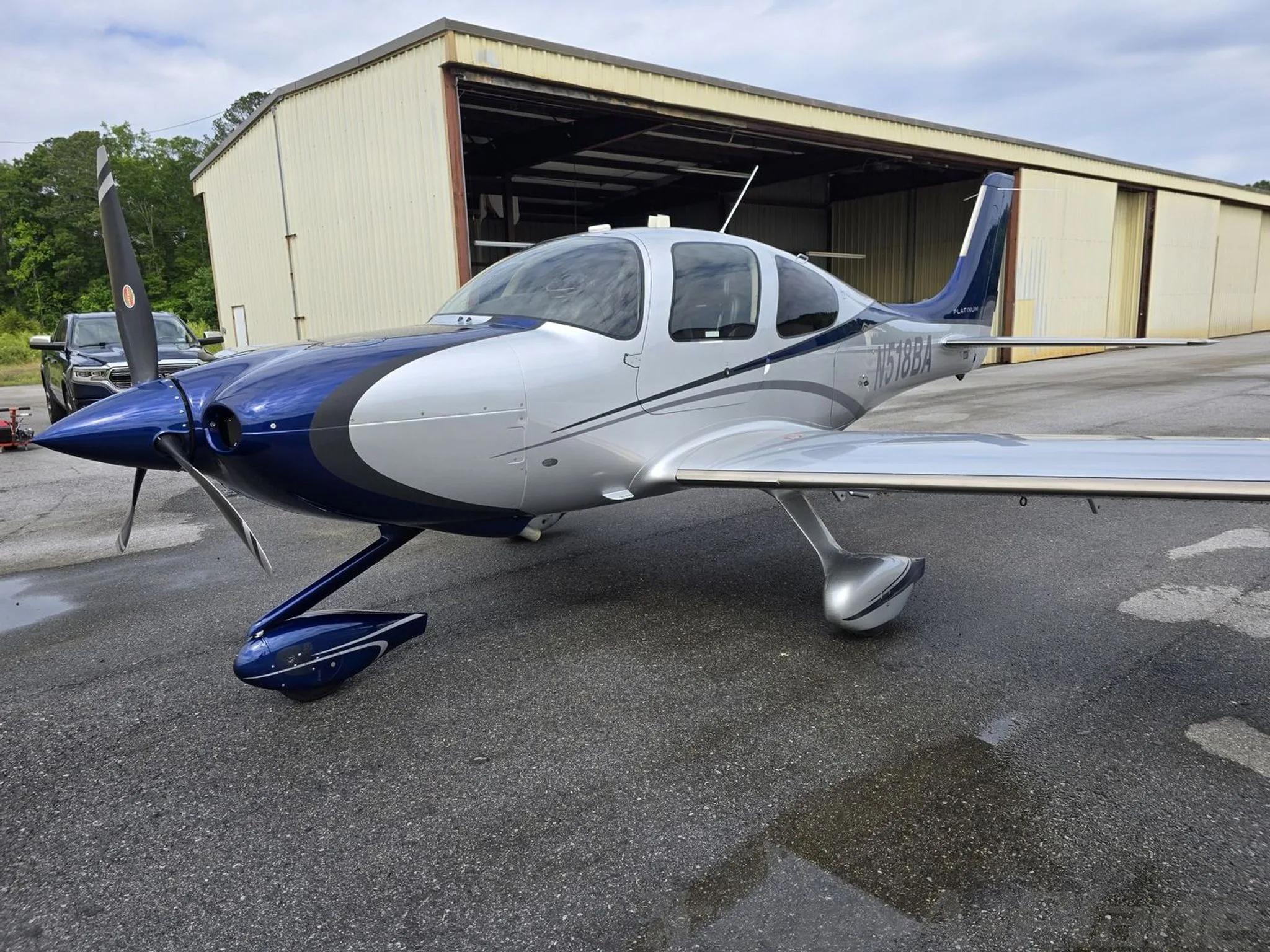 A blue and silver Cirrus SR22T aircraft, registration N518BA, parked on a wet tarmac in front of a hangar.