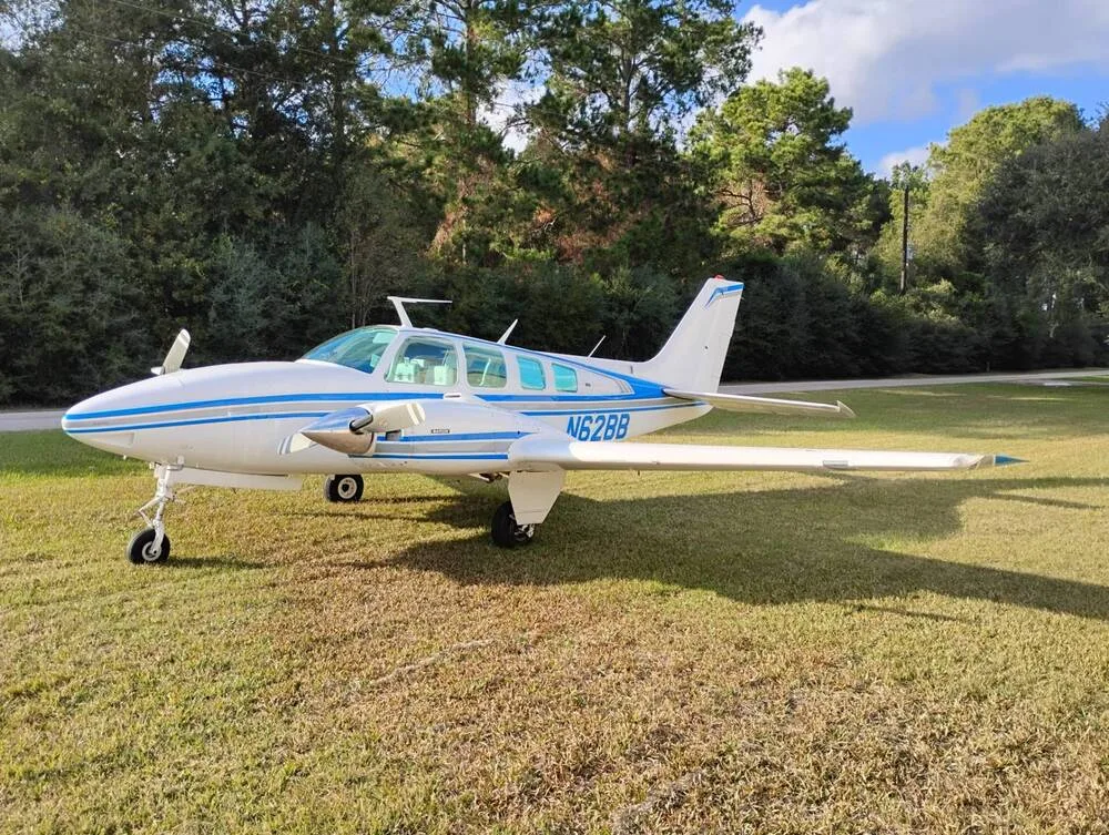 A white Beechcraft Baron N62BB with blue stripes is parked on a grassy field, viewed from the front-left.