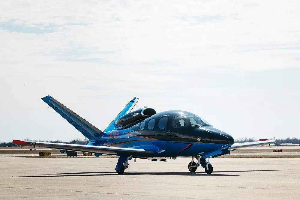 A Cirrus Vision Jet (N3EE) in blue and black paint scheme, parked on an airfield under a cloudy sky.
