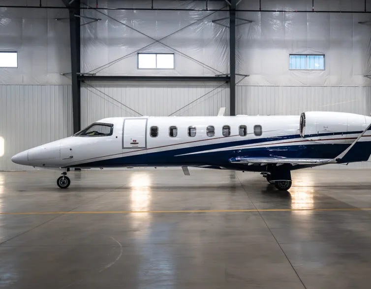 A white business jet with a blue stripe is parked inside a hangar, viewed from the side.