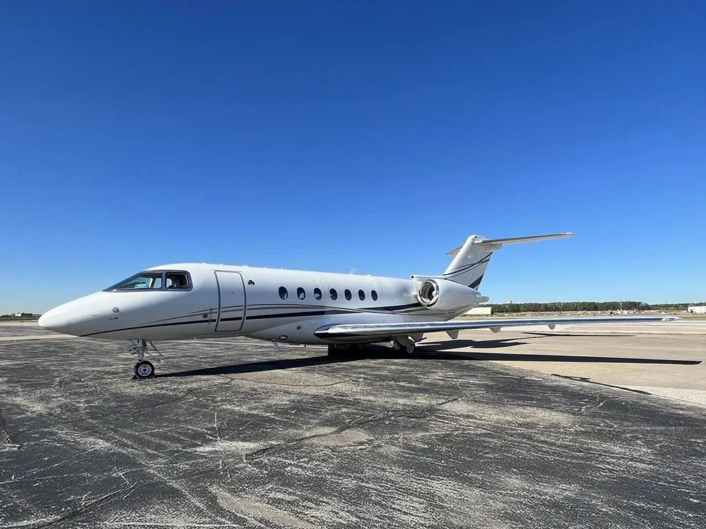 A white business jet with dark blue stripes is parked on an asphalt tarmac under a clear blue sky.