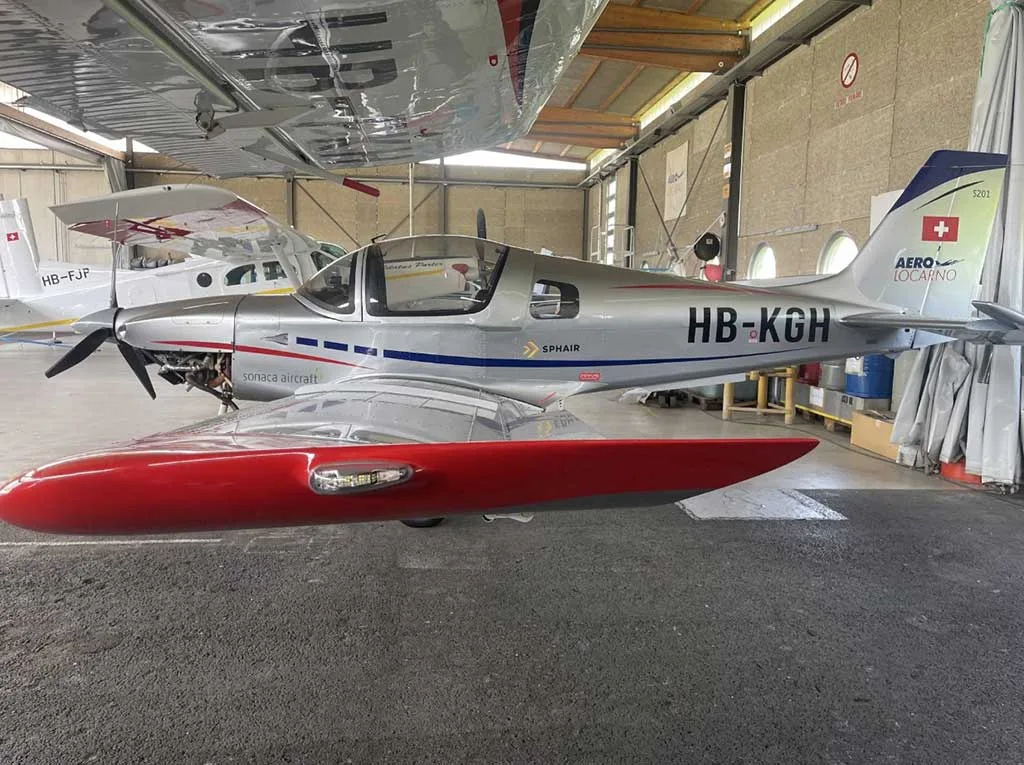 Side view of a silver Sonaca 200 aircraft with red wingtips and blue tail stripe, registration HB-KGH, parked in a hangar.