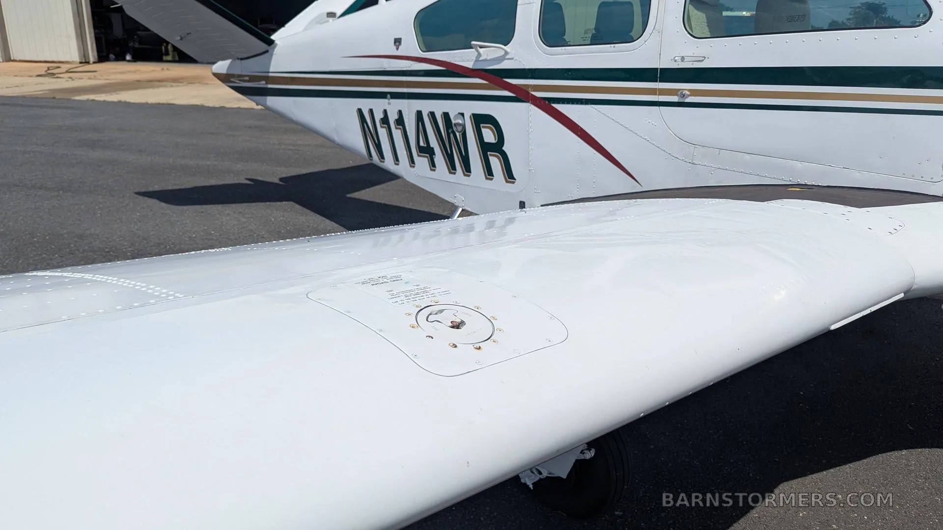 A close-up view of the white wing of the Beechcraft Bonanza, showing the fuel cap and part of the fuselage with N114WR registration.