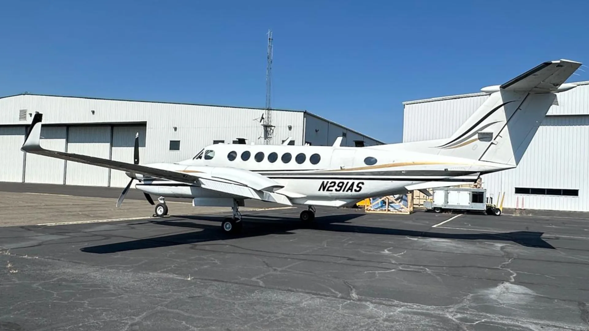 A white Beechcraft King Air N291AS with black and gold stripes parked on an asphalt tarmac in front of a hangar.