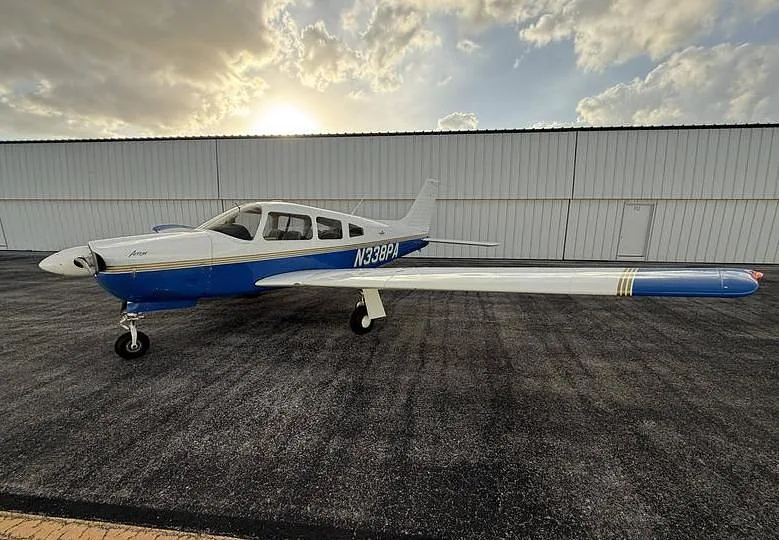 A white and blue Piper Arrow aircraft with registration N338PA parked on an asphalt tarmac in front of a hangar at sunset.
