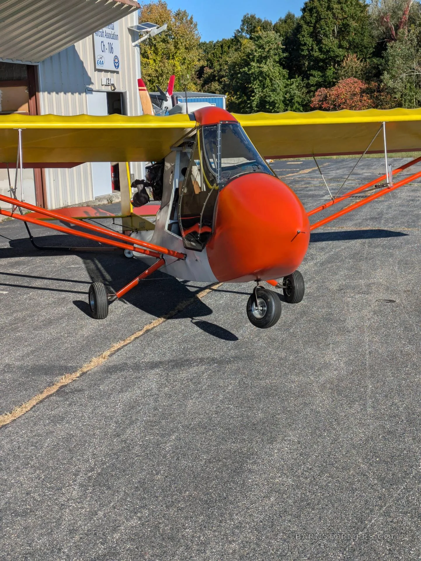 A front-quarter view of a red and yellow biplane-style ultralight aircraft parked on asphalt next to a hangar.