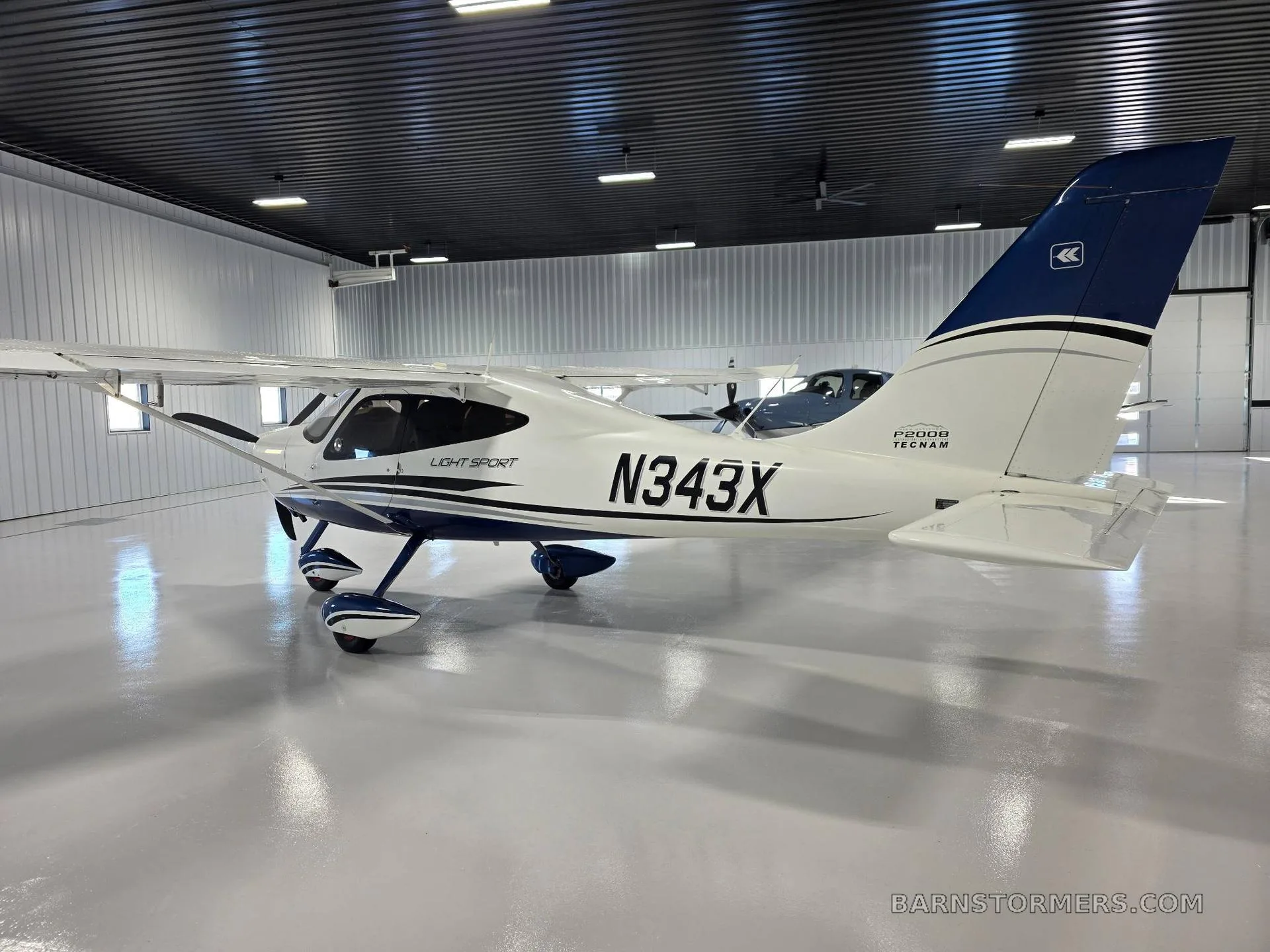 A rear-side view of a white Tecnam P2008 Light Sport aircraft with blue tail and accents, registration N343X, inside a hangar.