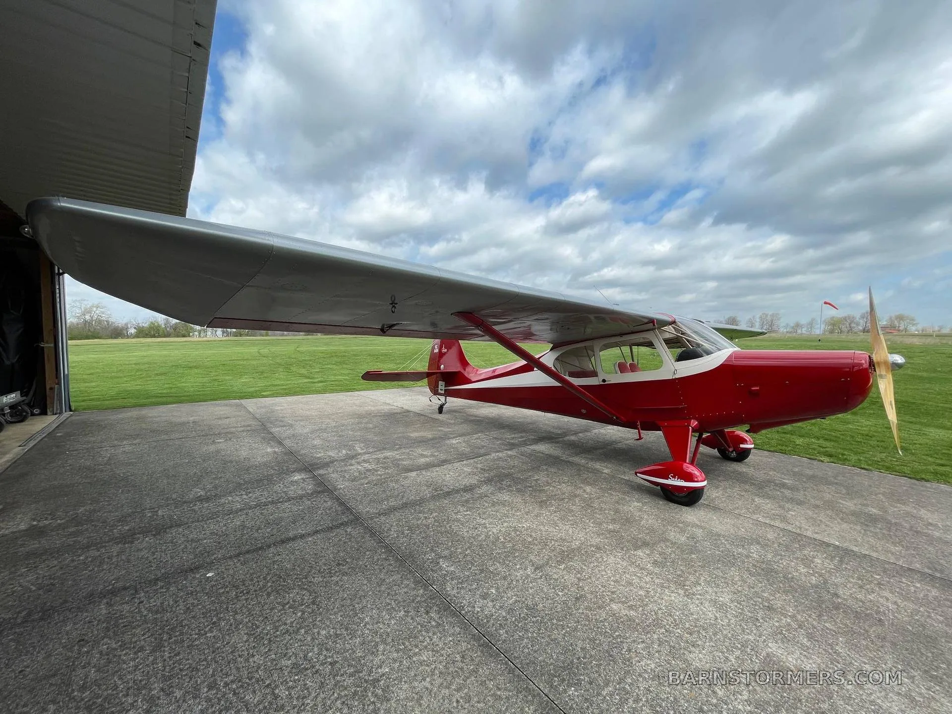 A red and white single-engine propeller aircraft with a high wing, parked on concrete next to a hangar under a cloudy sky.