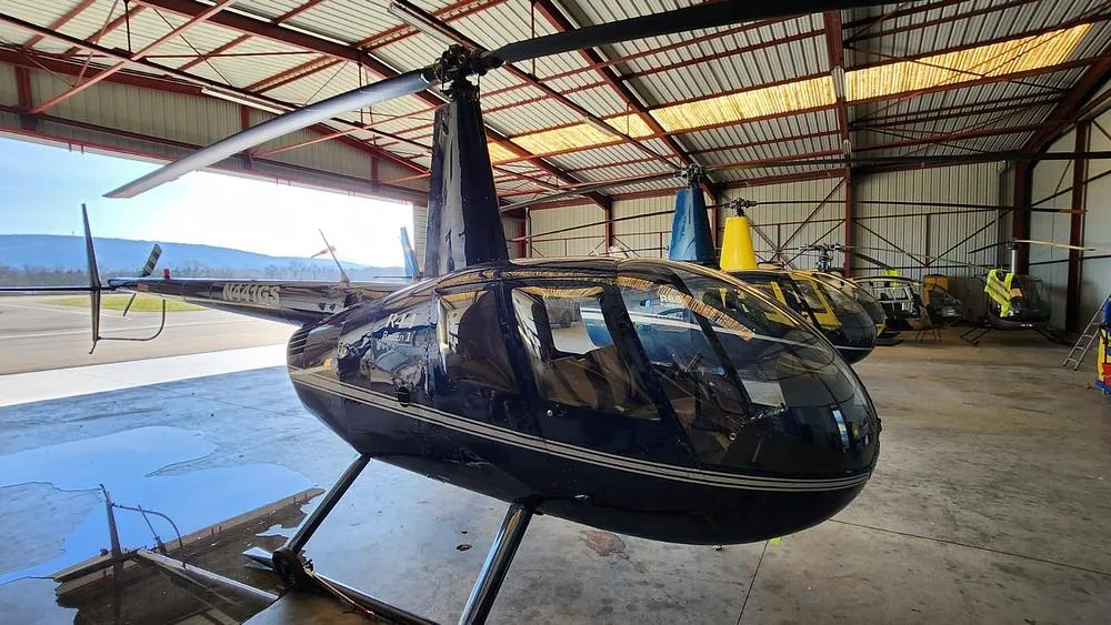 A dark blue Robinson R44 helicopter with white stripes parked in a hangar, viewed from the front-left.