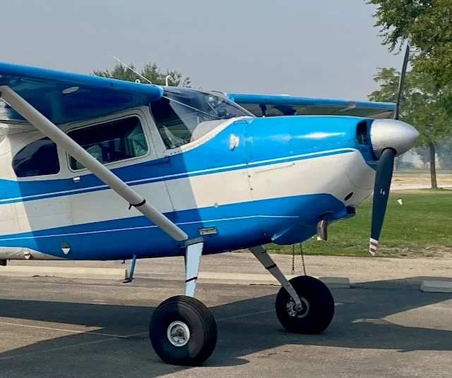 A blue and white high-wing propeller aircraft with large tires on a tarmac with trees in the background.