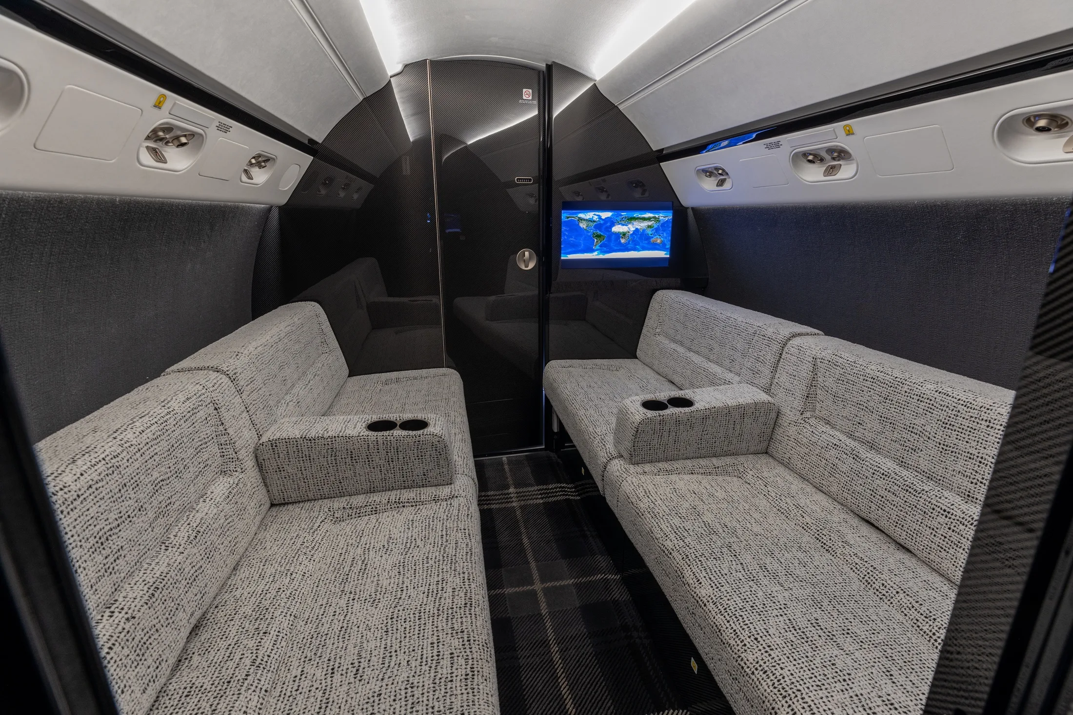 A private jet cabin section with two grey sofas facing each other, a dark plaid carpet, and a screen displaying a world map.