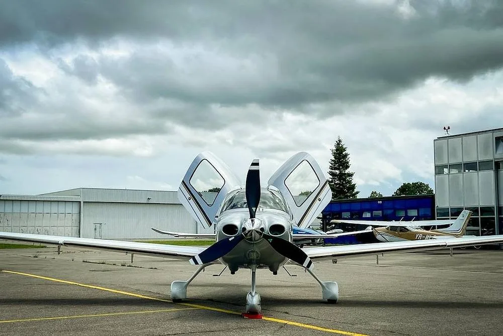 A front view of a white and silver Cirrus SR22 aircraft with its gull-wing doors open, parked on an airfield under a cloudy sky.