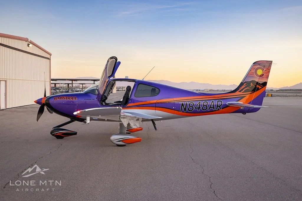 Side view of a purple and silver Cirrus SR22T with open doors, featuring a Colorado flag tail design, parked in front of hangars at sunset.