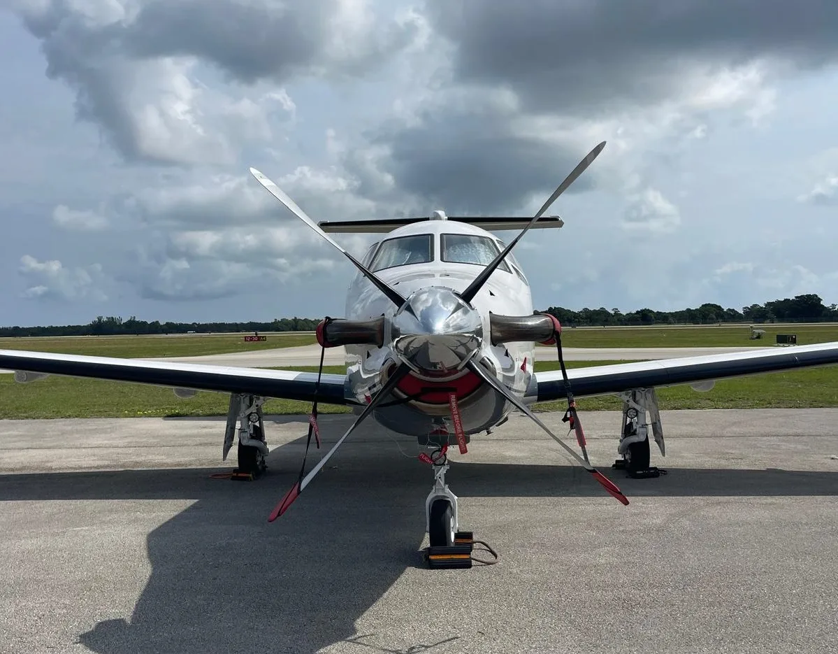 A head-on view of a white Pilatus PC-12 NG aircraft with blue and grey stripes, registration N542NG, parked on a tarmac.